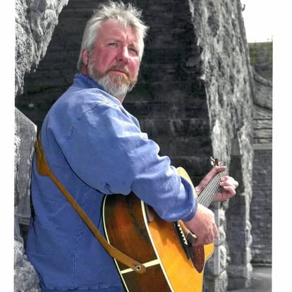 man holding a guitar while leaning against a stone wall